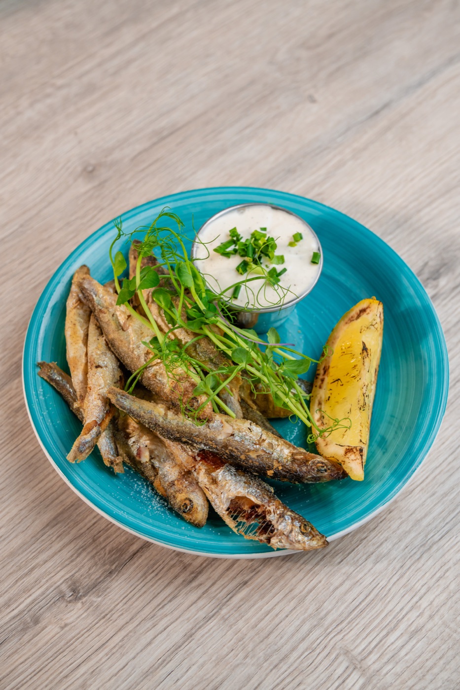 Szprotki bałtyckie smażone — podane na turkusowym talerzu z białym sosem czosnkowym ze szczypiorkiem, grillowaną cytryną i mikrozieleniną
